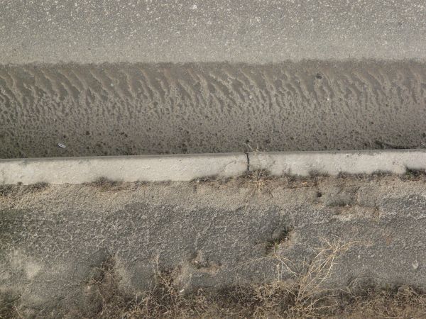 Road edge texture, with grey and white asphalt separated from a cracked sidewalk by a pattern left by melted snow. Brown weeds and fine dust cover the surface of the sidewalk.